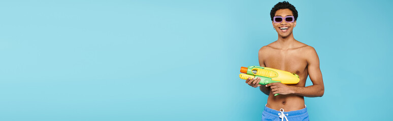 Naklejka premium Handsome young man enjoying a vibrant summer day with a water gun by a bright blue backdrop