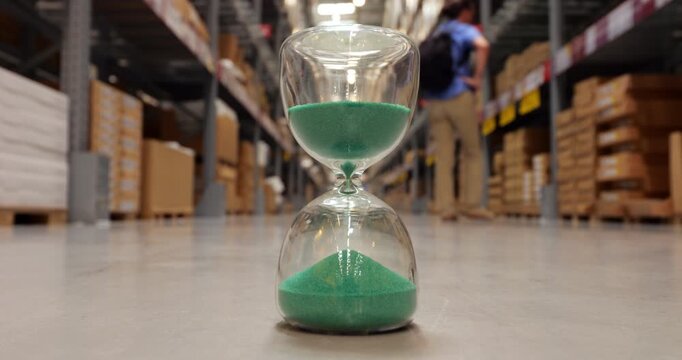Focus on hourglass with green sand standing on floor of large store warehouse, measuring passage of time while male customer walks in blurred background along shelves filled with boxes.