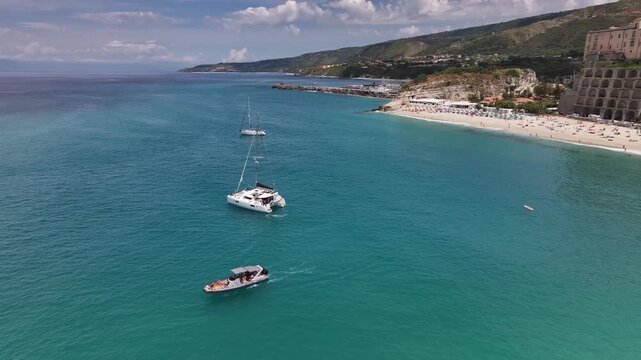 Drone pulling away from a catamaran while rotating to reveal a wide view of Tropea bay, coastline and historic town in Calabria, Italy.