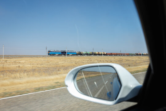 Long freight train passing through dry arid steppe landscape seen through car side mirror on empty highway in remote region