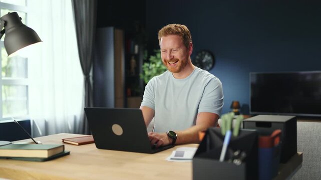A man sits at a desk in his home office. He types on his laptop and smiles. There is a lamp on the desk and a plant nearby. Sunlight comes through the window.