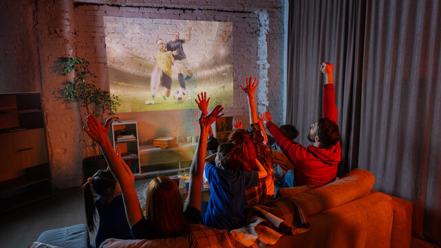 Family celebrating goal while watching soccer game projection in cozy living room. Concept of sports broadcast, football party, streaming service, home cinema, fan emotions, and family weekend.