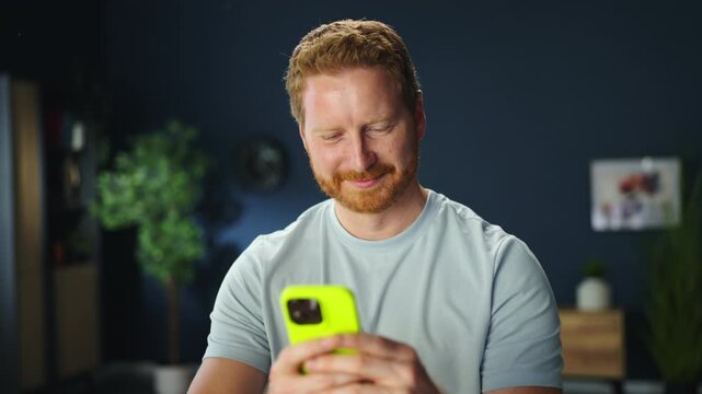 A man sits at his desk in a home office. He is looking at his smartphone while seated. A laptop and some papers are on the desk.