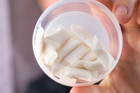 closeup of a hand holding an open round white plastic container filled with small nicotine pouches or snus bags in a bright indoor environment