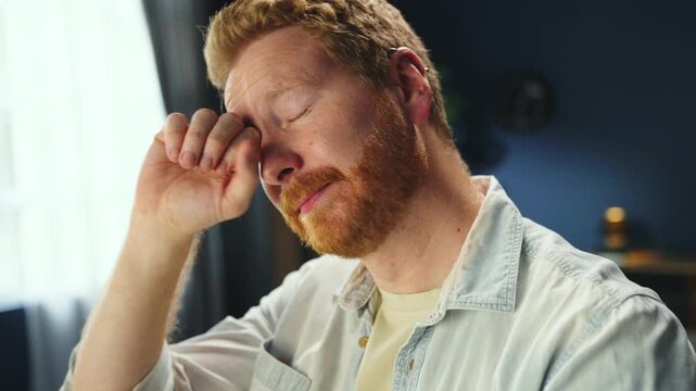 A man sits at his desk and yawns while looking at his computer. He appears tired and is trying to stay awake during work. The light from the window is bright.