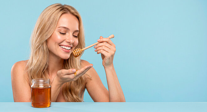 Beautiful blonde woman tasting bee honey on plain blue background. copy space. World honey bee day.