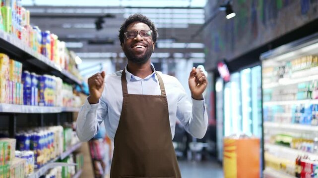 Happy cheerful african american supermarket salesman dancing between rows of shelves in market grocery store. Joyful funny seller worker in apron rejoices at workplace at marketplace. Dance of joy