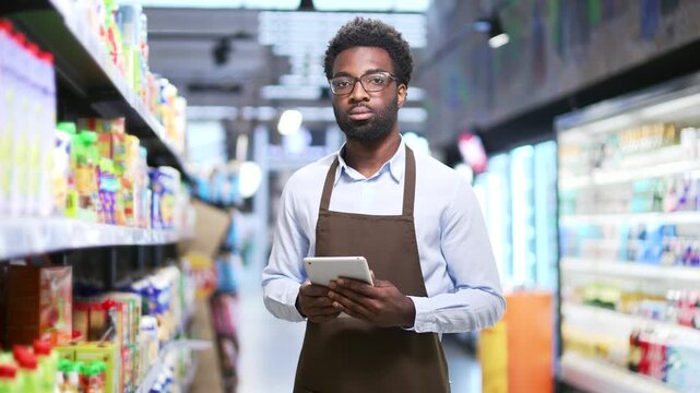Portrait of a serious African American supermarket worker standing between rows of shelves in market grocery store using tablet. Confident sales manager small business owner in apron looking at camera