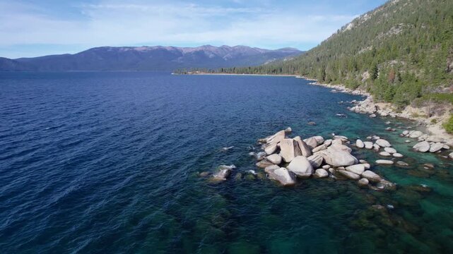 Cinematic aerial shot skimming the water surface with visible rock formations and alpine shoreline.