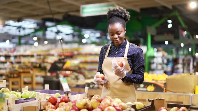 African american female greengrocer worker in supermarket checking condition of apples and picks in fruit department in market. Employee or small business owner working behind counter in grocery store
