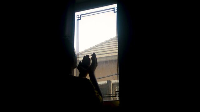 Man looking out window praying or contemplating during a rainy day with overcast sky and roof visible