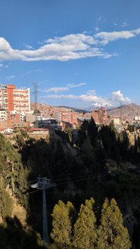 Scenic panoramic view of La Paz city center with green trees, Mi Teleferico infrastructure towers and residential houses on hills under blue cloudy sky with Illimani mountain background, Bolivia
