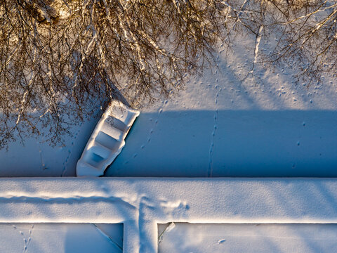 Aerial view of a snow-covered boat moored at a frozen pier with animal tracks on the ice and long shadows from bare trees in Trakai, Vilnius County, Lithuania.