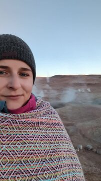 Happy young couple of tourists wearing traditional Andean ponchos and beanies taking a selfie at El Tatio geysers field during sunrise, winter adventure travel in Atacama desert, Chile