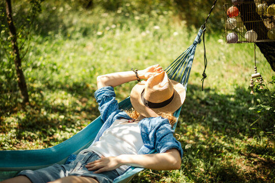Young woman lying in a hammock and covering her face with a straw hat while relaxing in a summer garden during holidays.