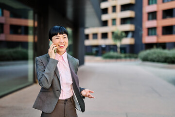 Fototapeta premium Portrait of a young asian japanese businesswoman woman using a smartphone mobile phone walking down the street, surrounded by moder corporate office buildings