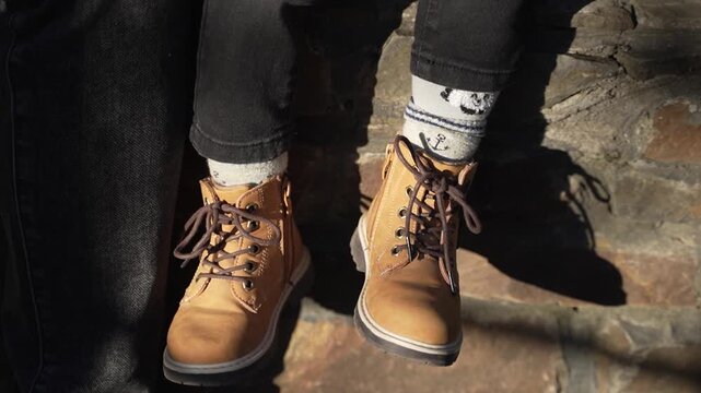 Child sits on bench in sunny autumn weather, dangling his legs in beige lace-up boots.