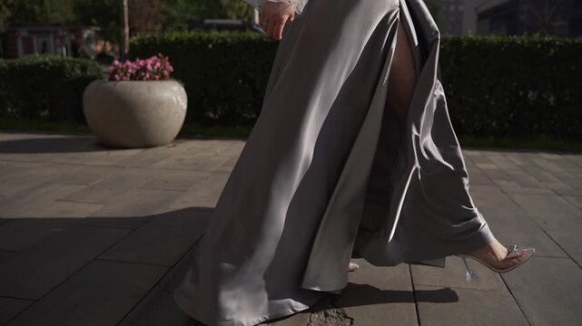 Young woman with beautiful slender legs walks down the street in silver dress with slit in slow motion. Bride in gray dress walks around the city during the day