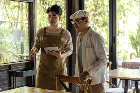Small business cafe owner guiding young barista, father and son discussing table setup while holding tablet, small business workflow planning and service improvement in cozy restaurant and coffee shop