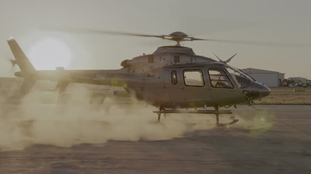 Helicopter lifts off tarmac at sunset. Smoke billows from landing gear. Sunlight casts warm glow on scene. Clear sky above, buildings in distance. Shadow of helicopter stretches across pavement.