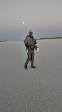 Happy male tourist walking and spinning under the full moon at twilight in Salar de Uyuni, smiling man enjoying freedom in Bolivia salt flats desert wearing winter clothes and chullo hat
