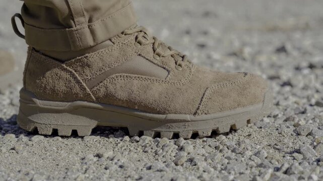 Tactical boots stand gravelly ground. Rugged soles grip rough terrain. Beige suede material covers shoe. Tan laces secure footwear tightly. Outdoor setting suggests military use. Sand-colored fabric.