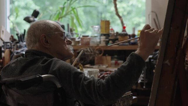 Senior artist on a wheelchair paints an oil painting on canvas in a studio