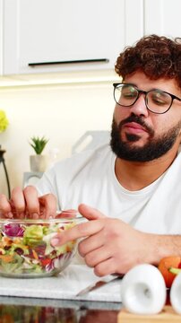 Arabian young man at kitchen table with new salad bowl looks tired and bored after strict keto diet. He forces healthy food but has no appetite and dreams of comfort cheat meals at home tonight again