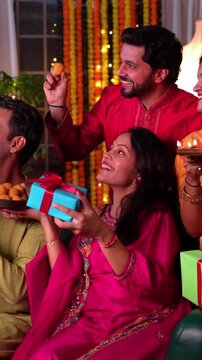  A beautiful happy Indian family of four in colourful festive wear bonding and capturing memories of Diwali celebration by lighting oil diyas, exchanging gifts and sweets in decorated living room