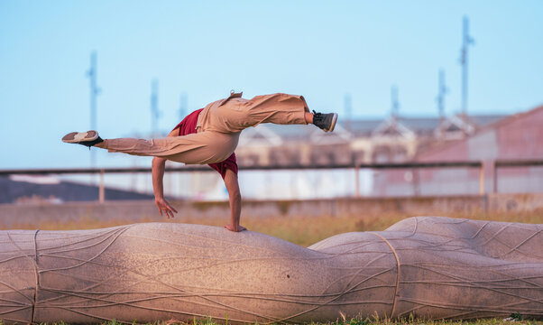 A female parkour athlete performs a dynamic one-handed handstand and leg split on a stone sculpture in an outdoor urban environment.