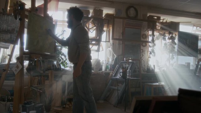 Anguished male artist drawing on canvas, then getting furious and destroying his painting during nervous breakdown in messy workshop filled with sunlight streaming through windows