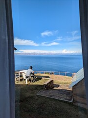 Fototapeta premium Man sitting on a patio looking out at a stunning deep blue lake framed by window curtains, peaceful travel and luxury relaxation concept at a scenic mountain resort in South America