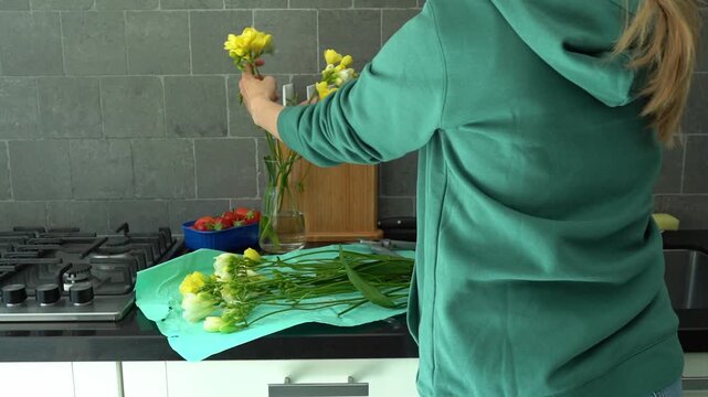 A woman in a green sweater trims fresh freesia flowers and arranges them in a vase on a wooden table, creating a bright floral centerpiece. Lifestyle blogs, gardening tutorials, florist workshop mater