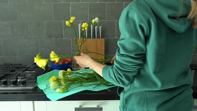 A woman in a green sweater trims fresh freesia flowers and arranges them in a vase on a wooden table, creating a bright floral centerpiece. Lifestyle blogs, gardening tutorials, florist workshop mater