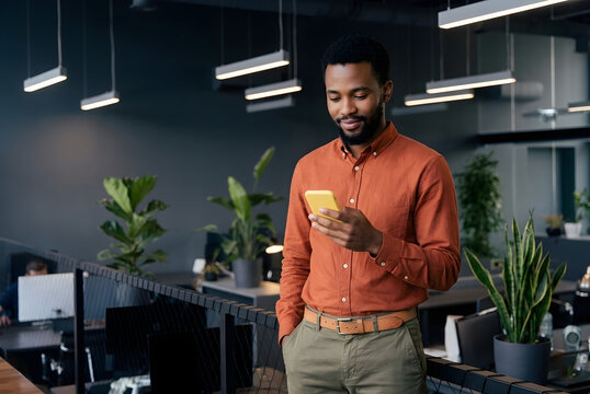 Business professional in modern office &ndash; Man in orange shirt with smartphone among desks, computers, and plants, highlighting workplace technology, digital communication, organizational privileges