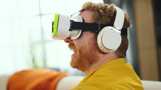 Man is using a virtual reality headset at home. He appears focused as he interacts with a virtual environment. The room is bright and comfortable, and he seems engaged in the experience.