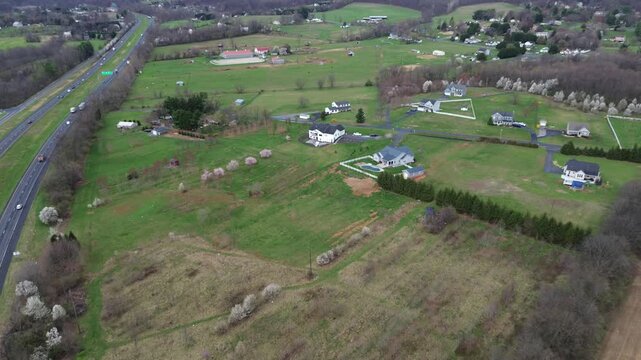 Semirural homestead cluster near Elmer Derr Rd with spaced residences, fenced yards, open grassy fields extending toward wooded pockets west of Frederick shows bright lawns, blooming trees, MD
