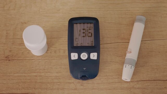 A glucometer sits on a wooden table showing a reading of 136 mg dL. Other diabetes tools are also on the table. Focus is on blood sugar measurement.
