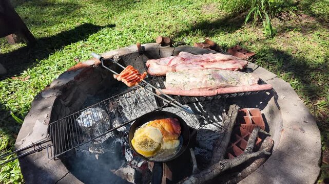 A variety of meats and bread cook over an open fire in a backyard setting. The scene captures the rustic charm of outdoor grilling. Suitable for culinary videos or cultural documentaries.