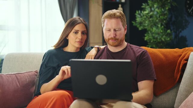 A couple sits on a couch in their living room. They engage with each other while using a laptop. The setting appears cozy with plants and soft lighting.