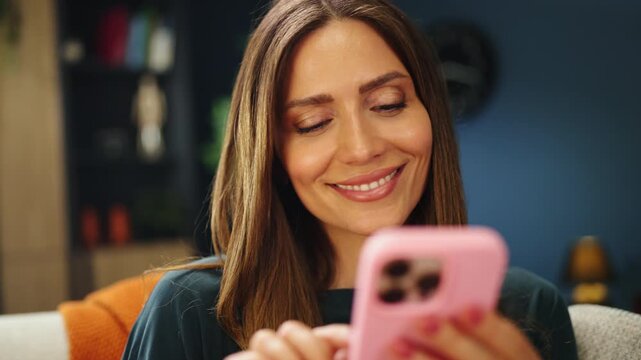 A woman sits on a couch and looks at her phone with a smile. She seems happy while interacting with her device in her living room during the daytime.