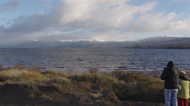 A couple stands by the shoreline of Huechulafquen Lake, gazing at the distant Lanin Volcano. The scene captures the expansive Patagonian landscape. Perfect for travel features.