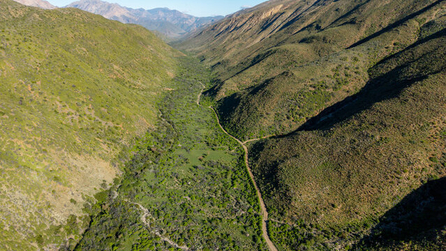Aerial view of Die Hel Gamkaskloof valley with a winding dirt road surrounded by steep green mountains under a clear sky in Central Karoo District Municipality, Western Cape, South Africa.