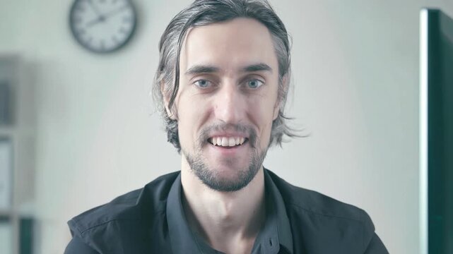 office crazy worker at workplace: Medium-length dark-haired man with facial hair sitting at office desk, calmly smiling with brief eye closure against softly lit neutral background with wall clock