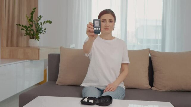A woman displays a glucometer reading of 200 mg dL while seated in her living room. This reading indicates a high glucose level that needs attention.