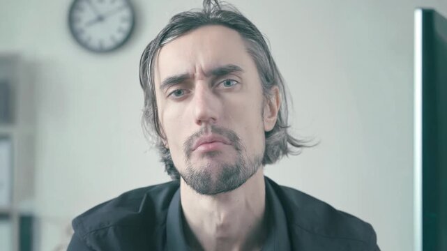 Bored office worker at workplace seated indoors against light background wearing black shirt with tousled hair, serious and contemplative with furrowed brows in soft neutral light and blurred clock