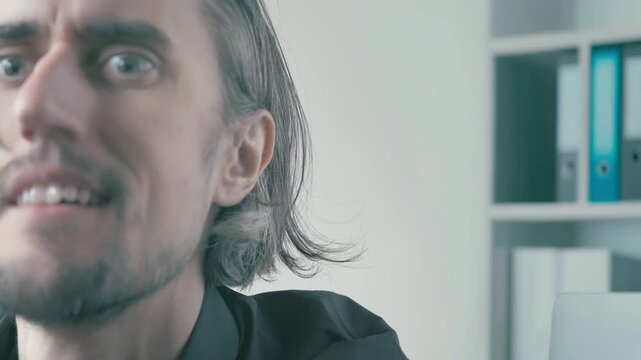 Crazy mad office worker at workplace silent went insane - Close-up of long dark hair man with facial hair in bright office lighting shifting from confused to warm broad smile looking toward camera