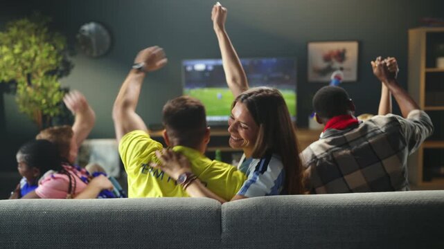 Friends sit together in a room and enjoy a soccer match on TV. They cheer and wave their arms as they celebrate plays. Drinks and snacks are present on the table.