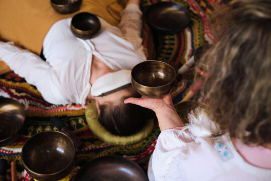 High angle view of a therapist playing a Tibetan singing bowl near a woman resting with an eye pillow. Private sound bath rituals support stress relief, deep relaxation, and emotional balance.