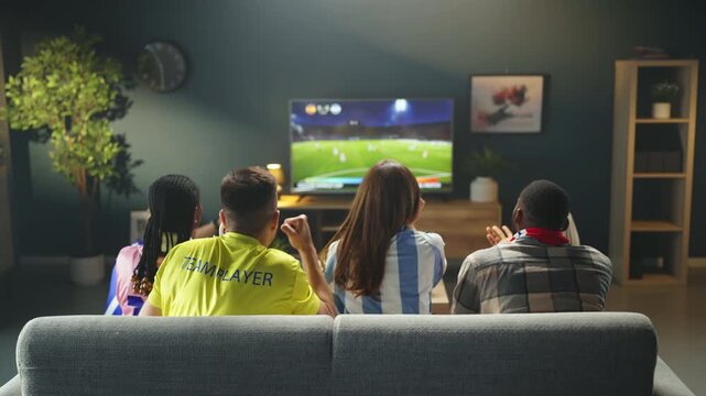 Groups of friends sit on a couch watching a sports game on TV. They cheer and react to plays while enjoying snacks. The setting is a cozy living room with some decorations.
